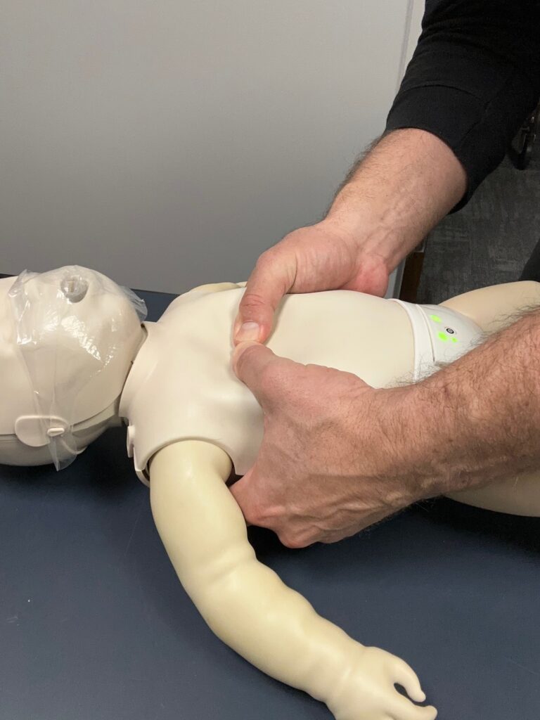 Two‑thumb infant CPR chest compressions on a baby manikin during AHA‑based CPR class in Atlanta, showing correct hand placement for parents and caregivers.